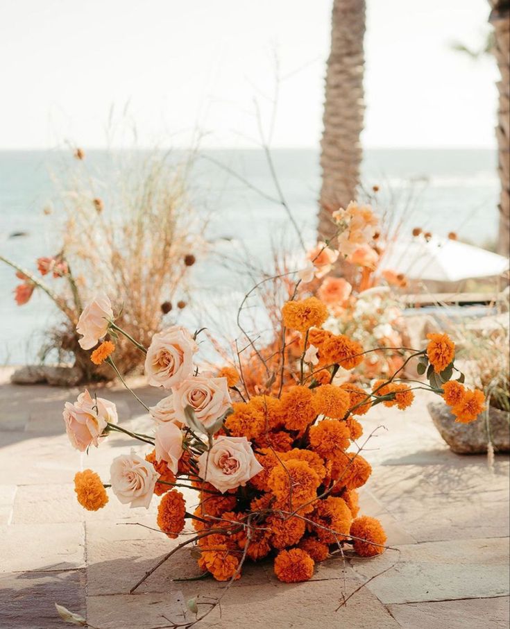 an arrangement of orange and white flowers sitting on the ground near some water with palm trees in the background