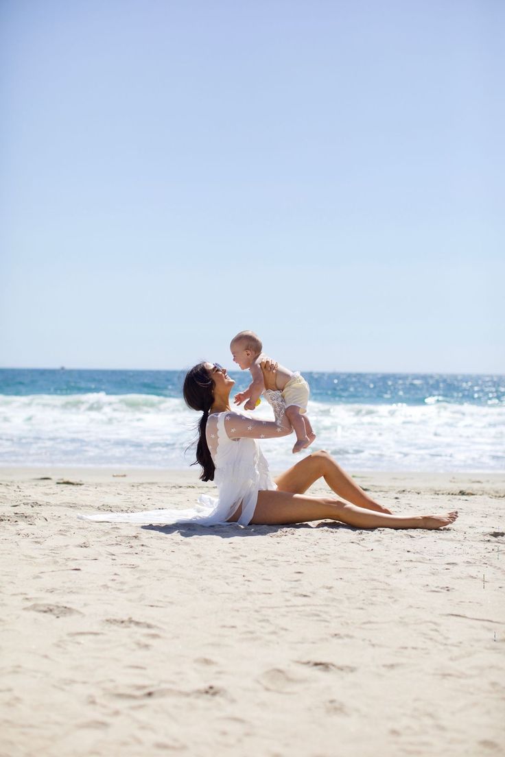 a woman holding a baby while sitting on top of a sandy beach next to the ocean