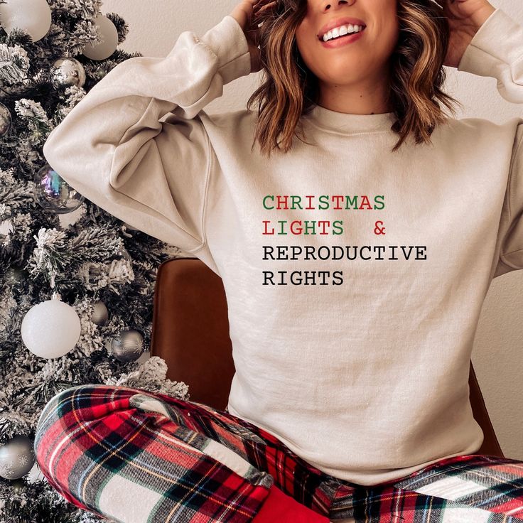 a woman sitting in front of a christmas tree wearing a merry sweatshirt and plaid pants