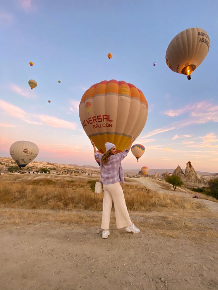 a woman standing on top of a dirt field with hot air balloons flying in the sky