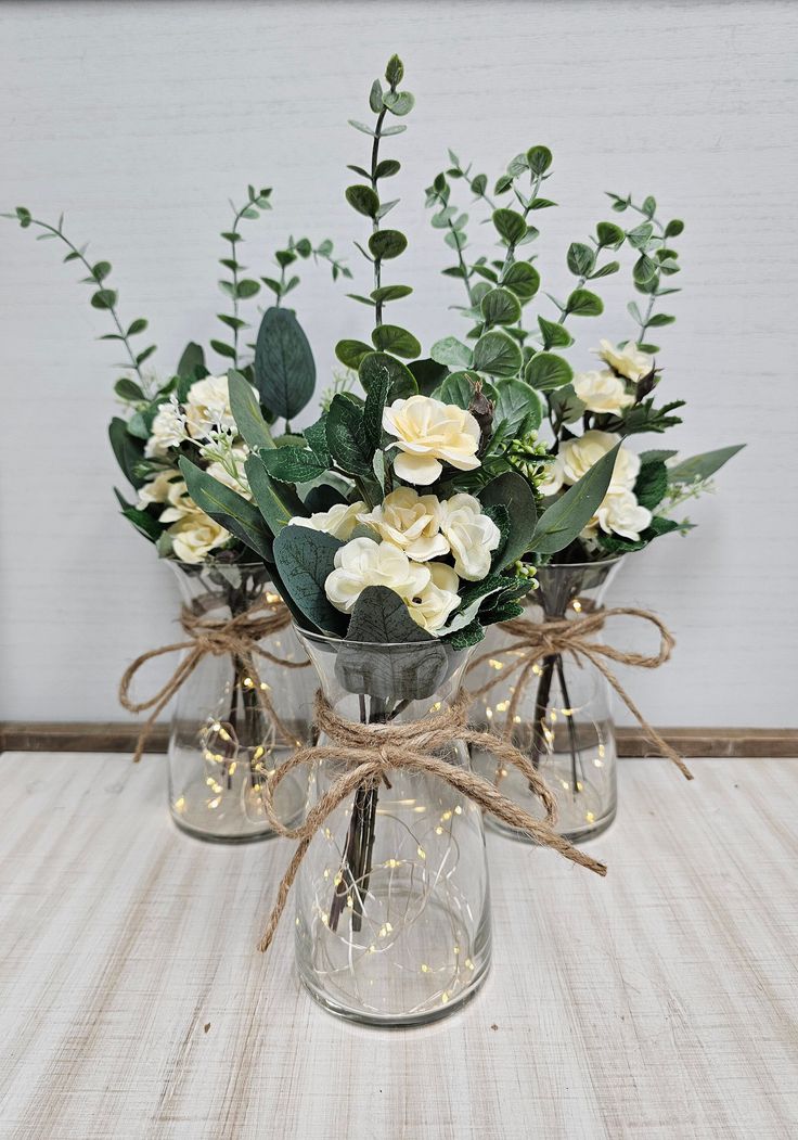 two clear vases with white flowers and greenery tied to each other on a table