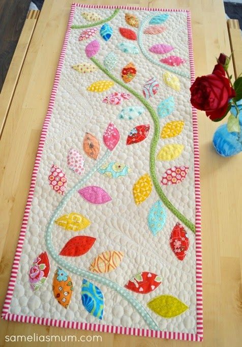 a quilted table runner with flowers and leaves on the edge, along with a vase filled with red roses