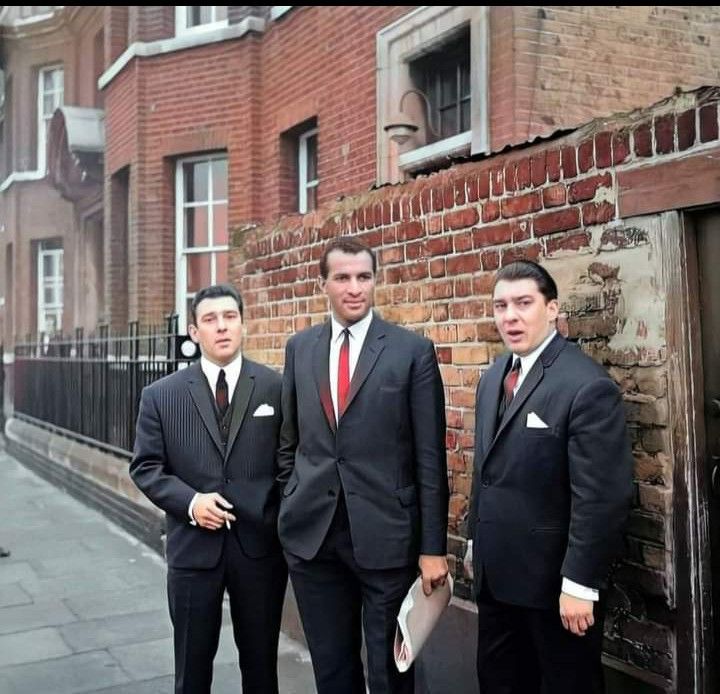 three men in suits standing next to each other on the side of a road near brick buildings