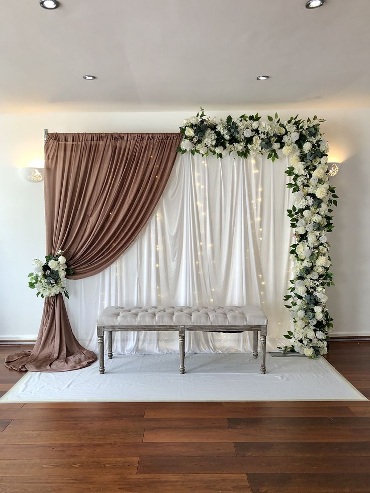 a wedding ceremony setup with white flowers and greenery on the wall, along with a bench