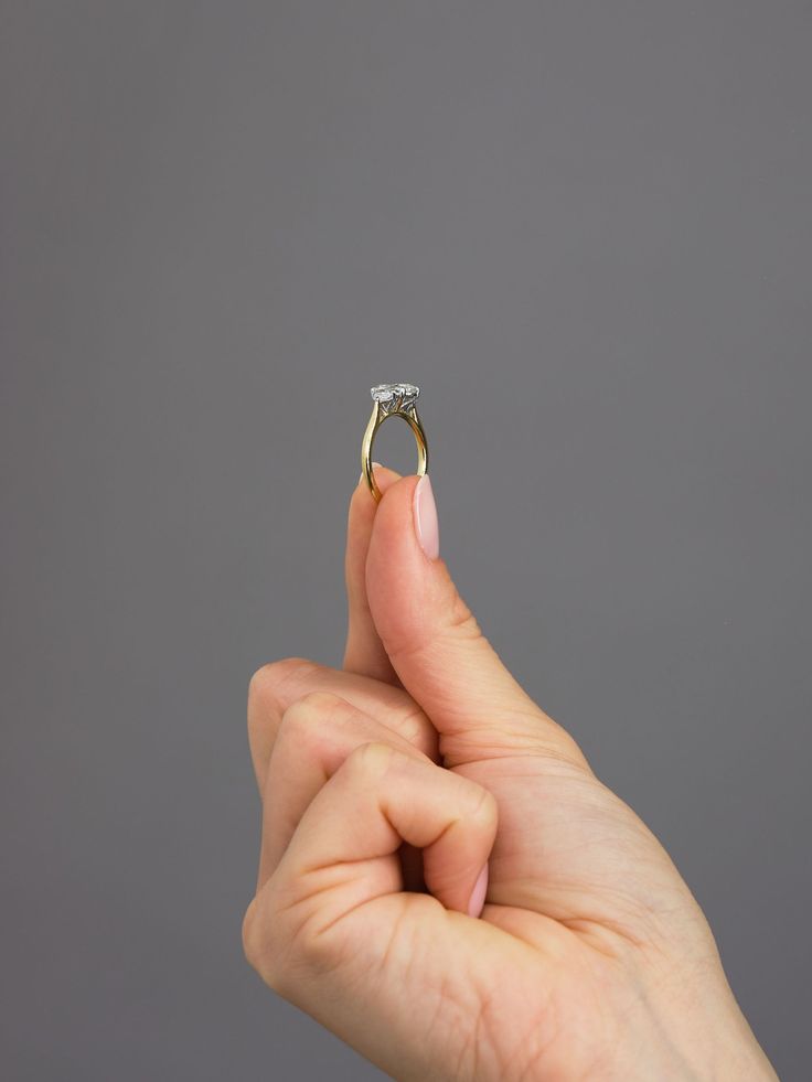 a hand holding a diamond ring in front of a gray background with the middle finger pointing towards it