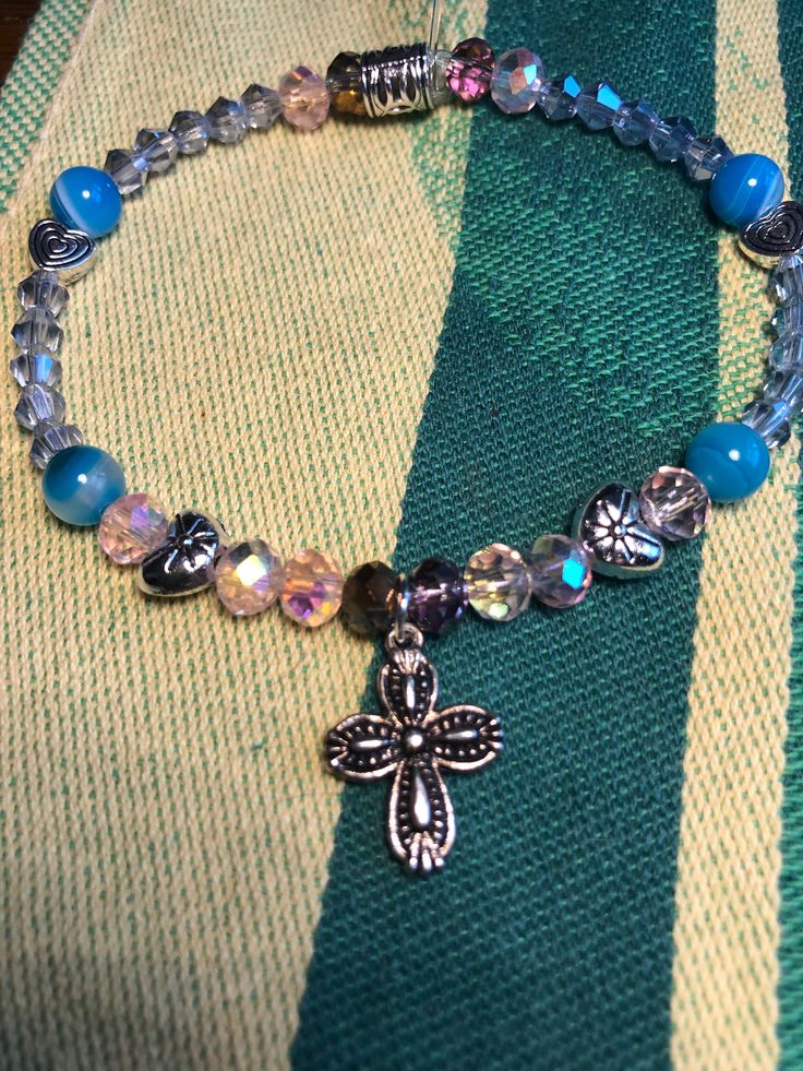 This silver-pated cross charm bracelet is filled with little surprises, like silver metal heart beads, light pink aurora borealis faceted glass beads, striped blue agate, and light blue glass beads. This bracelet is sized for a child. Silver Crystal Bracelet With Heart And Round Beads, Silver Crystal Bracelet With Heart Shaped Beads, Blue Charm Bracelet With Heart Beads And Adjustable Fit, Adjustable Blue Charm Bracelet With Heart Beads, Spiritual Silver Cross Charm Bracelet, Spiritual Silver Charm Bracelet With Cross, Blue Hypoallergenic Charm Bracelet With Round Beads, Adjustable Blue Rosary Bracelet With Cross, Hypoallergenic Blue Charm Bracelet With Round Beads