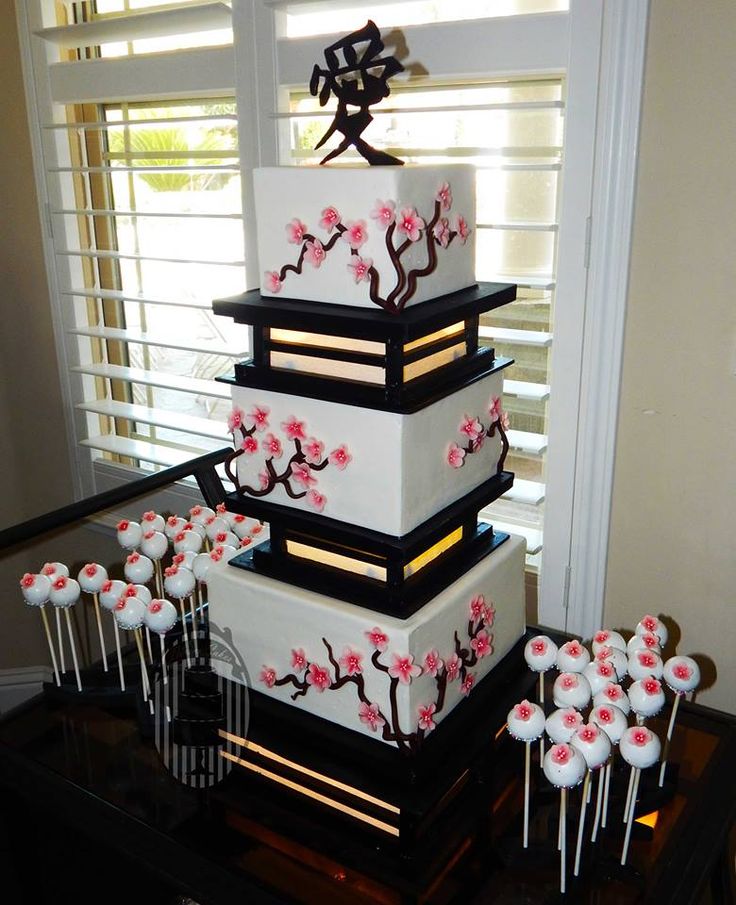 a white and black four tiered cake with pink flowers on the top, surrounded by small sticks