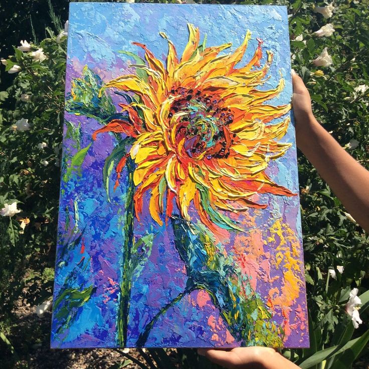 a painting of a sunflower is being held up by someone's hand in front of some flowers