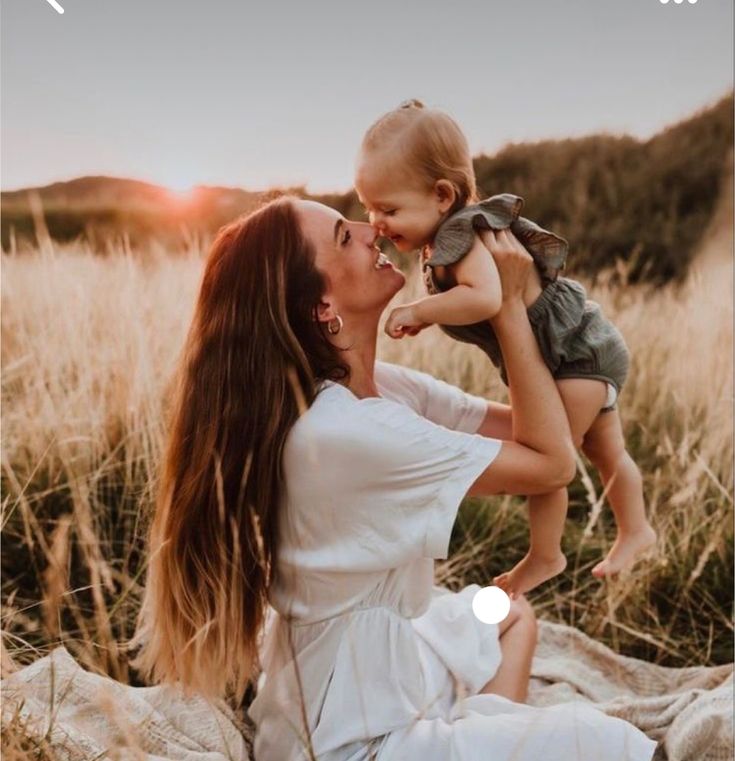 a woman holding a small child in her lap while sitting on a rock with the sun setting behind her