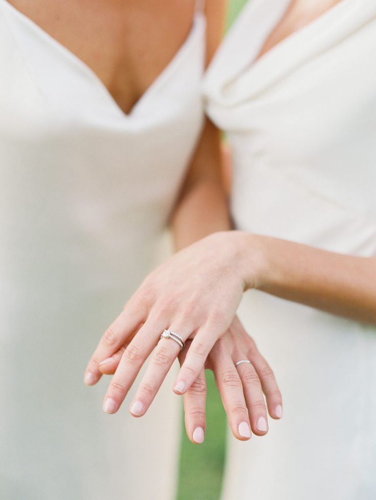 two women in white dresses with their hands on each other's shoulders, one holding her wedding ring