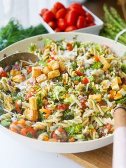 a white bowl filled with pasta salad next to tomatoes and broccoli on a table