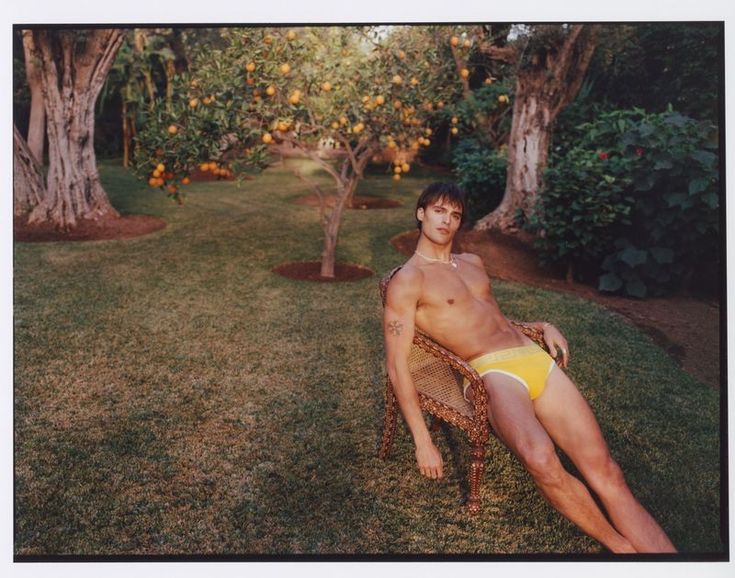 a shirtless man sitting on a chair in the grass near an apple tree grove