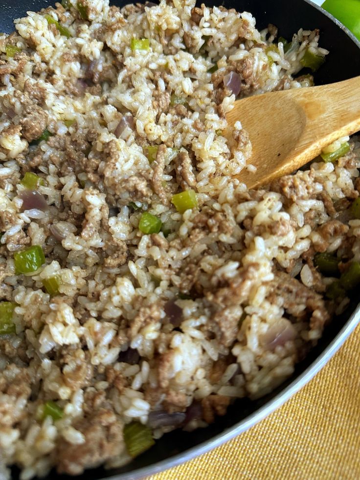 a skillet filled with rice and vegetables on top of a yellow cloth next to a wooden spoon