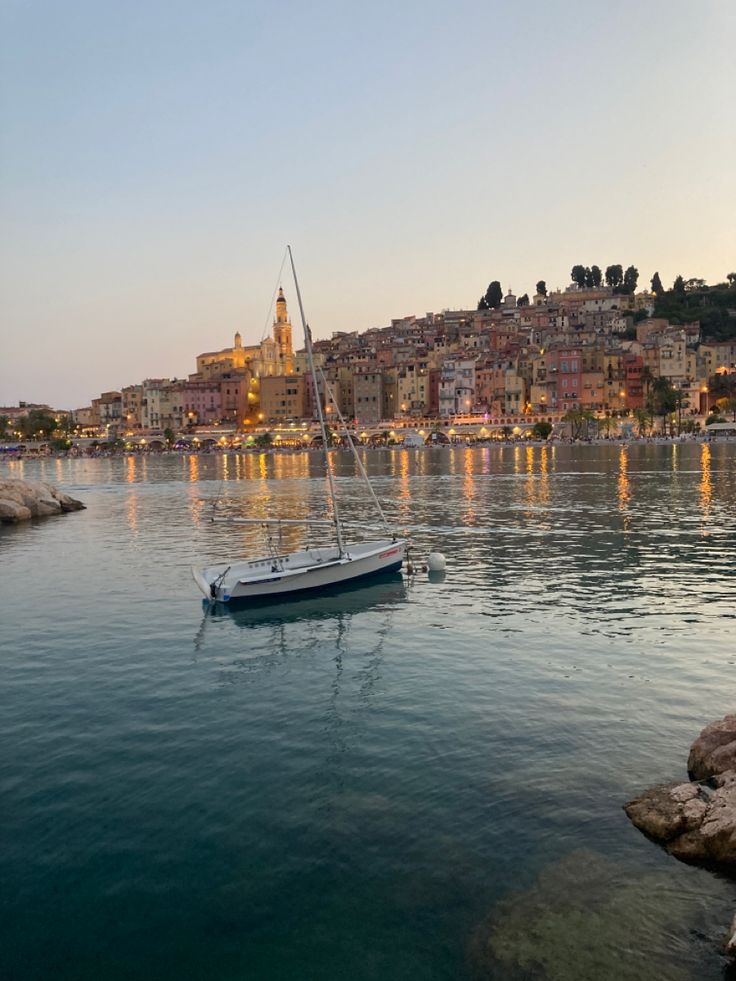 a sailboat floating on the water in front of a city at sunset or dawn