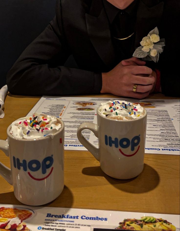 two coffee mugs with whipped cream and sprinkles on them sitting at a table