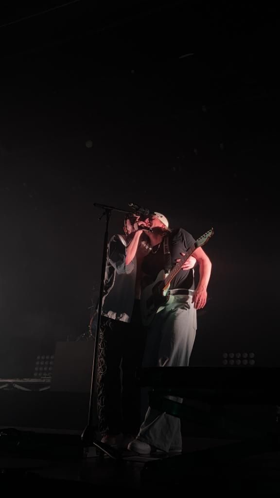 two men standing on stage with microphones in their hands and one holding his head to his ear