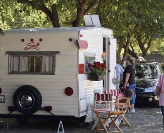 an old camper is parked next to some chairs and tables with flowers on them