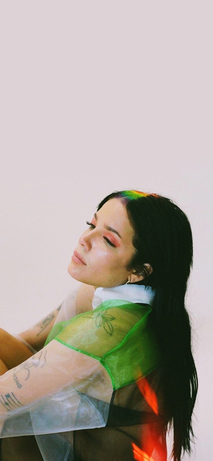 a woman with long black hair and tattoos on her arm, sitting in front of a white wall