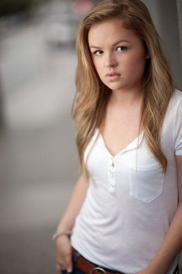 a woman with long blonde hair wearing a white shirt and jeans standing next to a wall