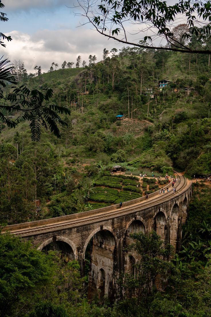 an old bridge in the middle of a lush green hillside with people walking on it