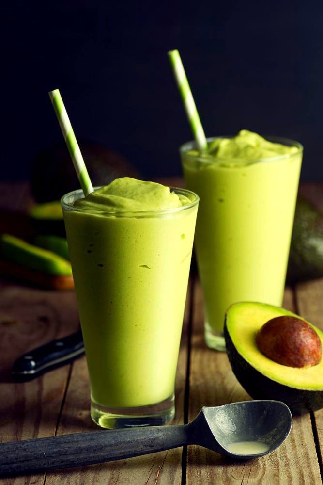 two glasses filled with green smoothie next to an avocado