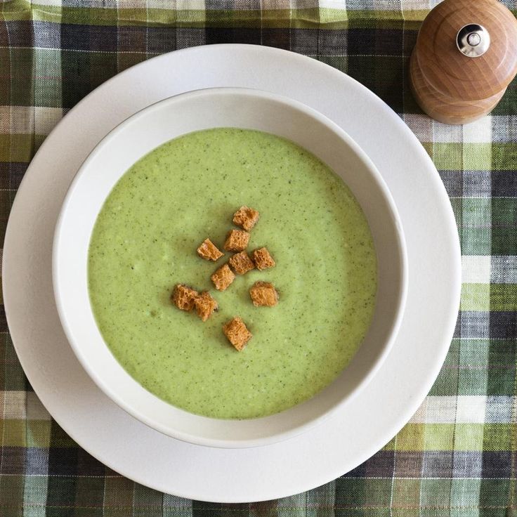 a white bowl filled with green soup and croutons on top of a checkered table cloth