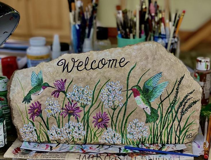 a welcome stone with flowers and hummingbirds painted on it, surrounded by paintbrushes