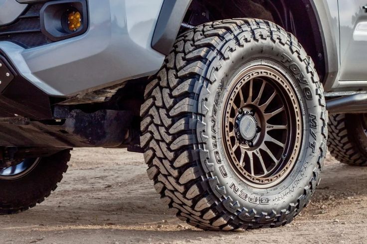 the front wheels and tires of a silver truck on dirt ground with rocks in the background