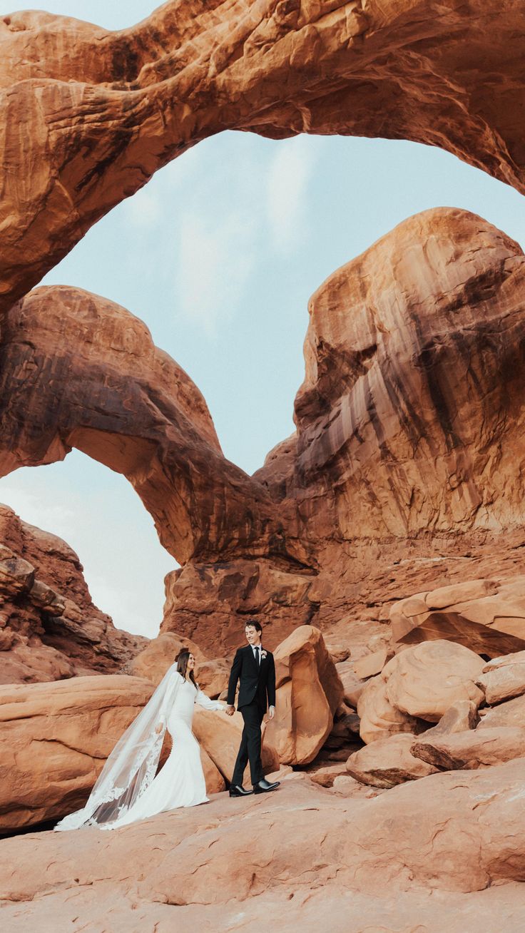 a bride and groom standing in the desert under an arch shaped rock formation at their wedding