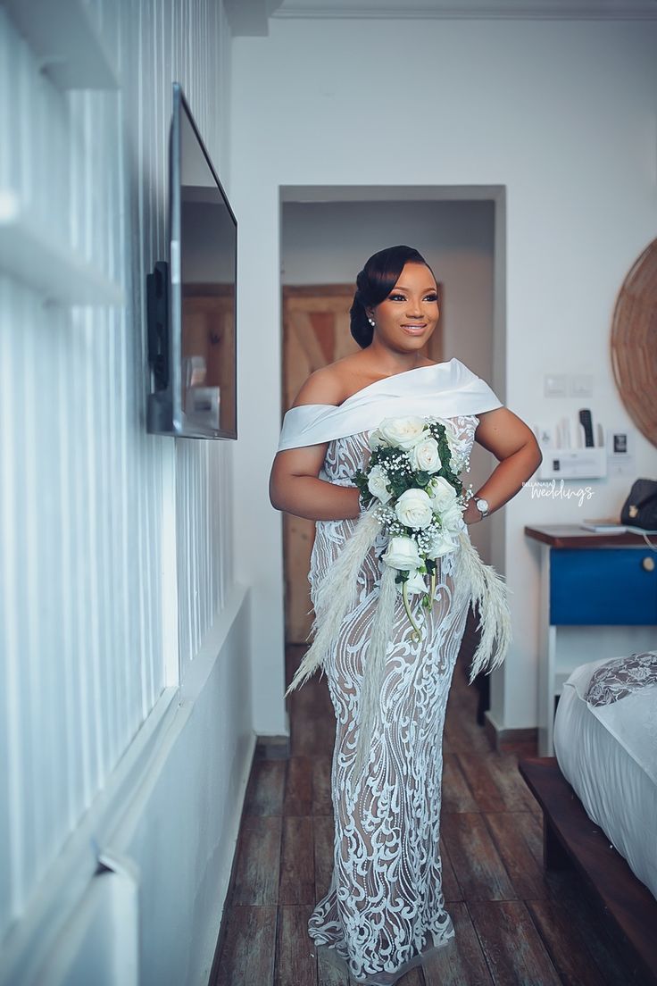 a woman in a white dress holding a bouquet and standing next to a bed with her hands on her hips