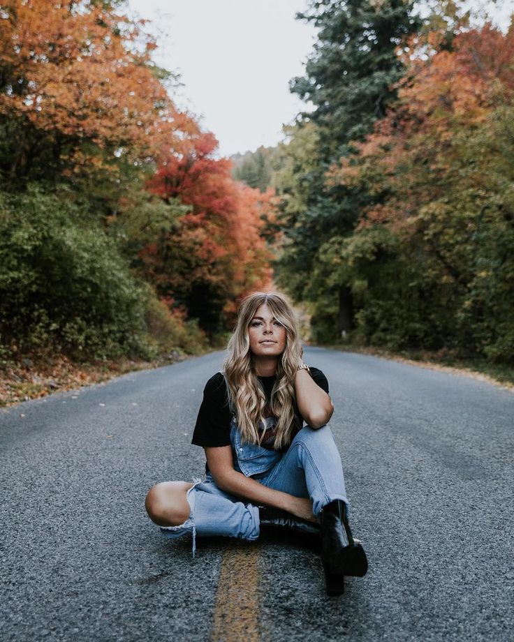 a woman sitting on the side of a road in front of trees with fall colors