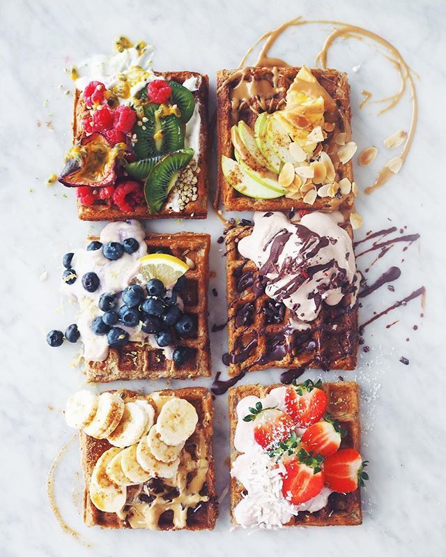 four waffles with different toppings are arranged on a marble surface, including bananas, strawberries, and blueberries