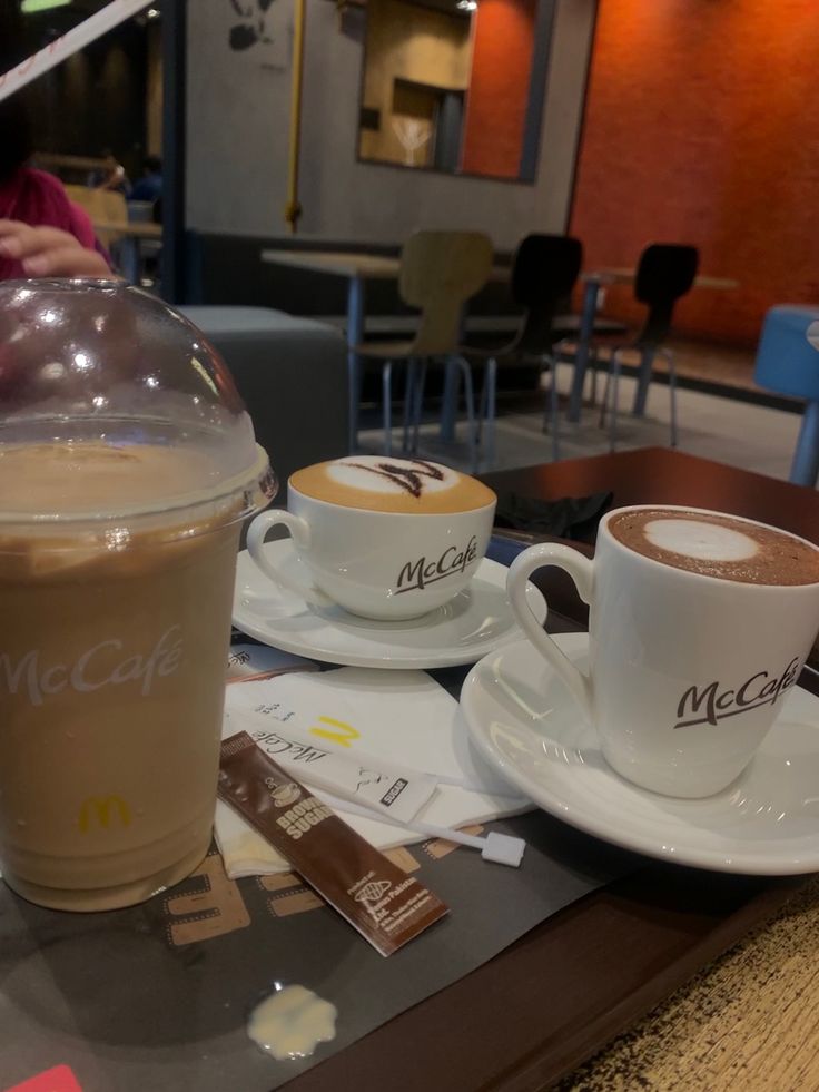 two cups of coffee sitting on top of a table next to each other with the word mcfc cafe written on them