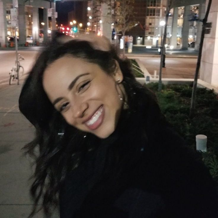 a woman is smiling while standing on the sidewalk in front of a building at night