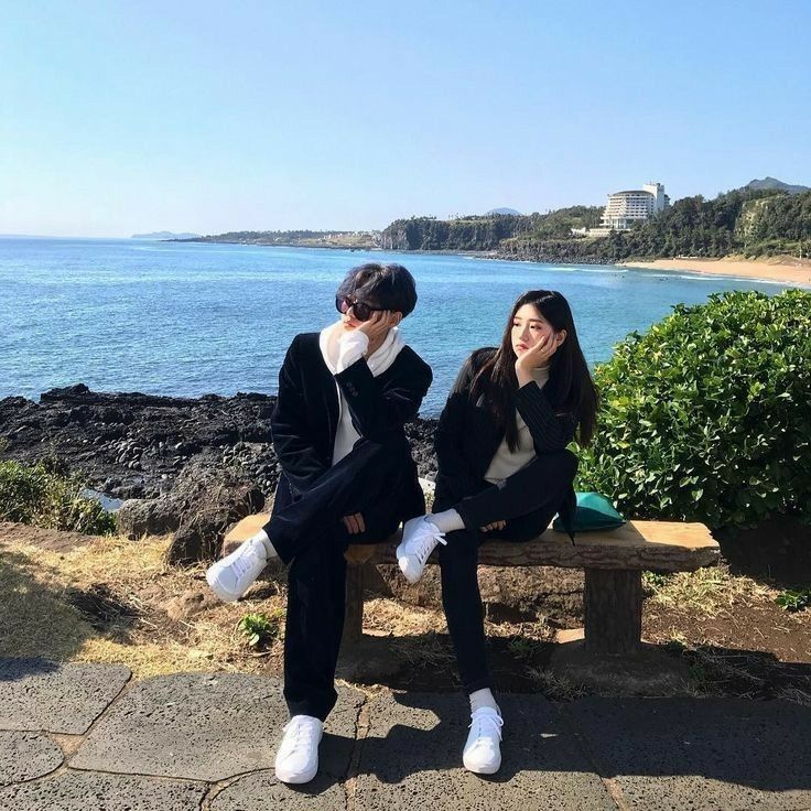 two young women sitting on a bench near the ocean with their feet up in the air