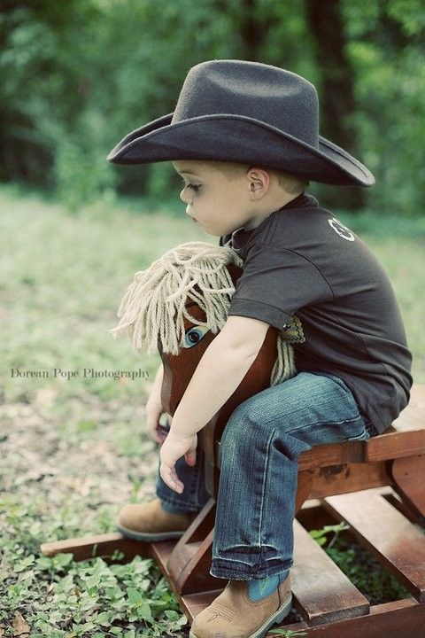 a young boy in a cowboy hat sitting on a wooden toy horse with grass and trees behind him