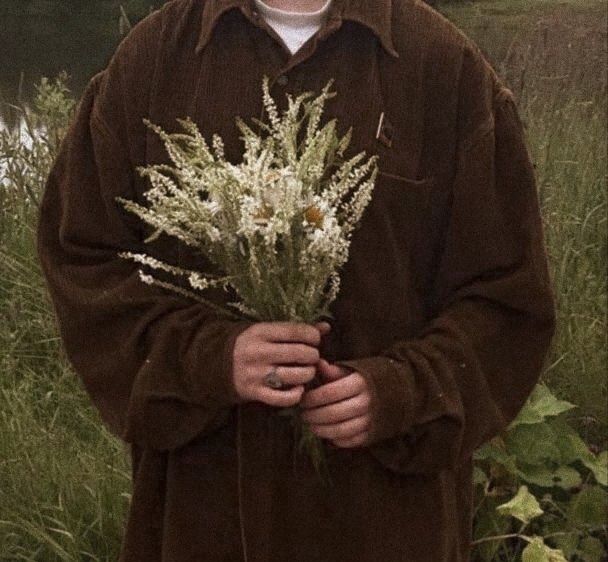 a man standing in the grass holding a bunch of white flowers on his chest and wearing a brown coat