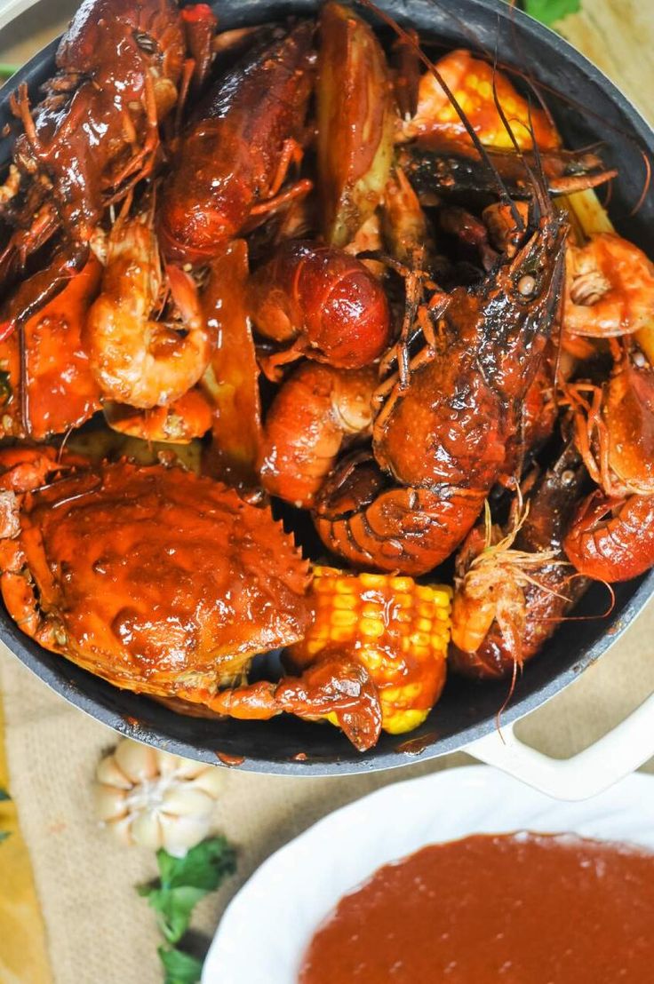 a pan filled with cooked lobsters and corn on the cob next to a bowl of sauce