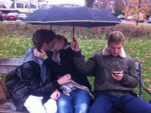 three people sitting on a bench with an umbrella over their heads and one person holding a cell phone