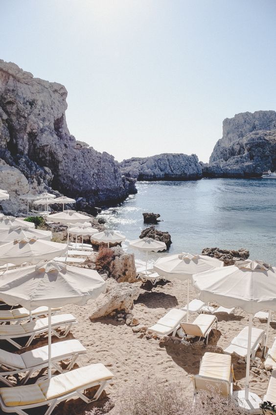 many chairs and umbrellas are on the beach near the water's edge, with rocky cliffs in the background