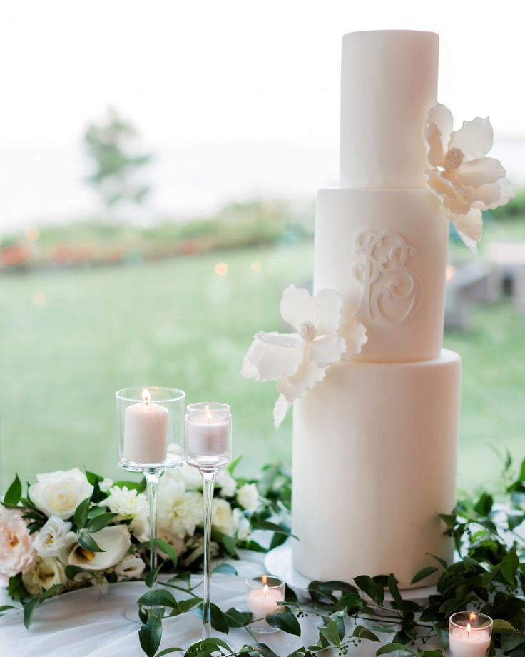 a white wedding cake sitting on top of a table next to candles and flowers in front of a window