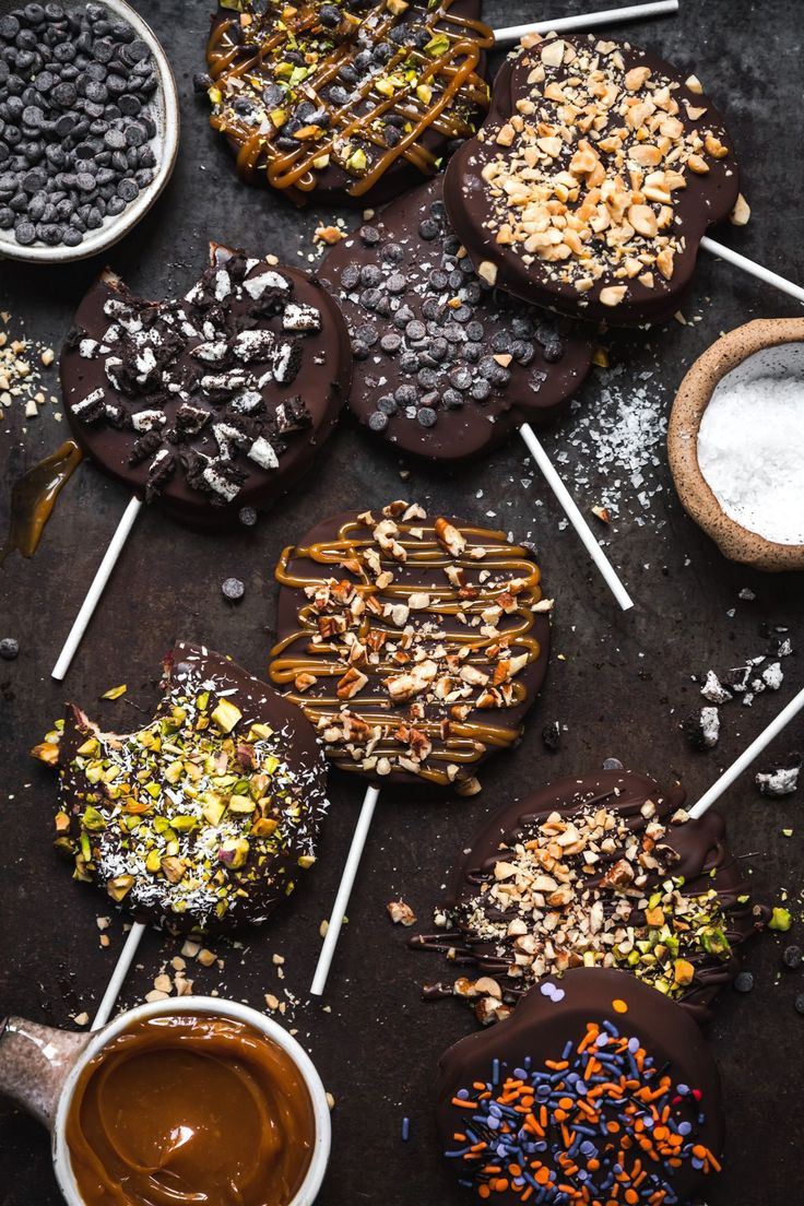 chocolate covered cookies with sprinkles and toppings on sticks next to bowls of confections