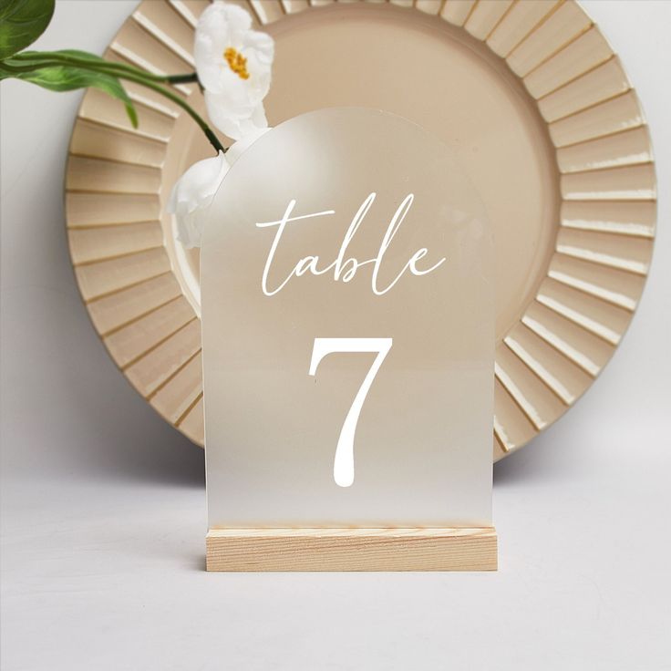 a table number with a white flower on it and a gold plate in the background