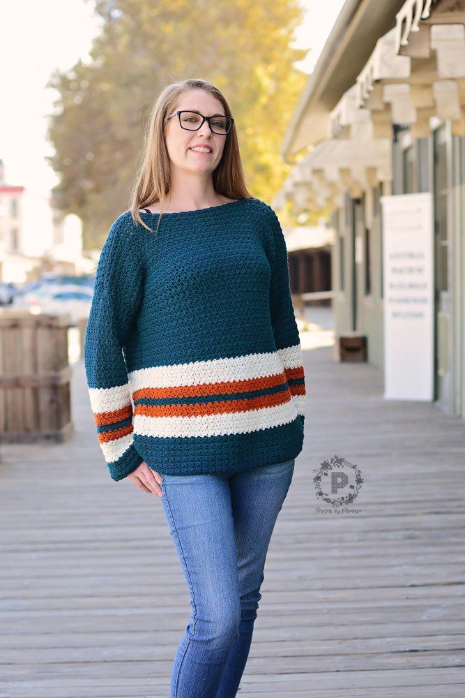 a woman standing on a wooden walkway wearing a green sweater and blue jeans with an orange, white, and red stripe