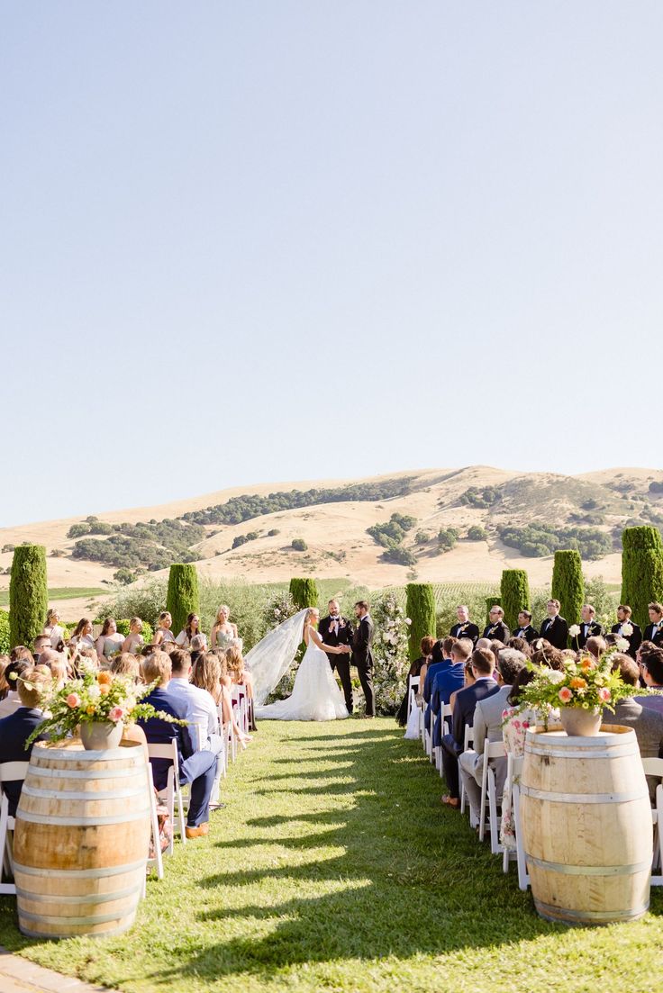 a wedding ceremony in the middle of an open field with wine barrels and greenery