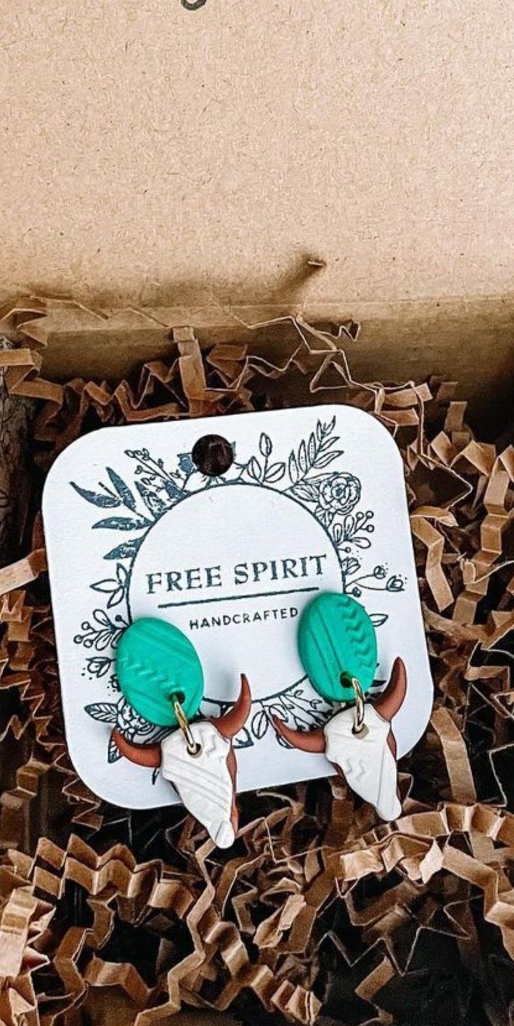 two green and white earrings sitting on top of a cardboard box filled with wood chips