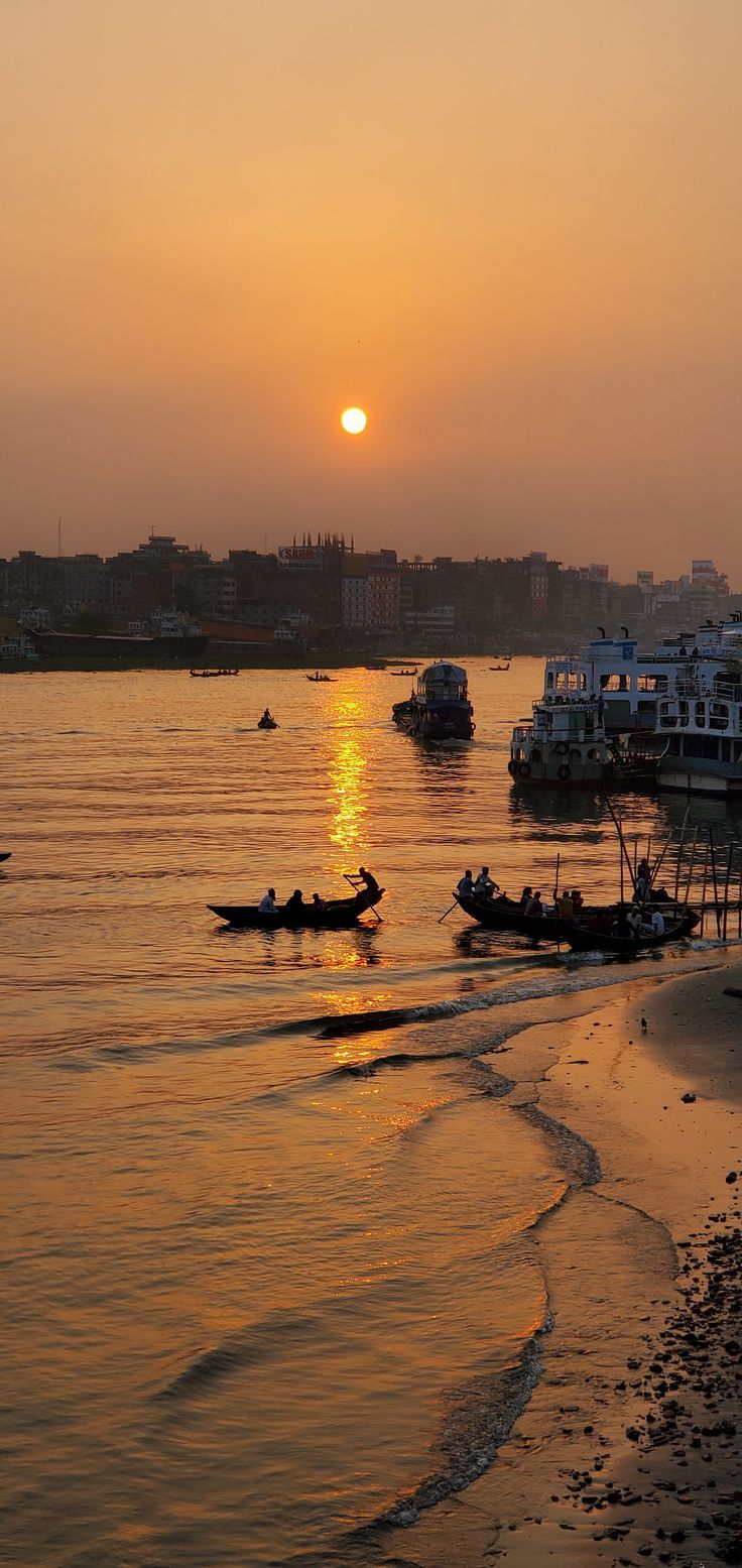 the sun is setting over some boats in the water on the beach with buildings behind them