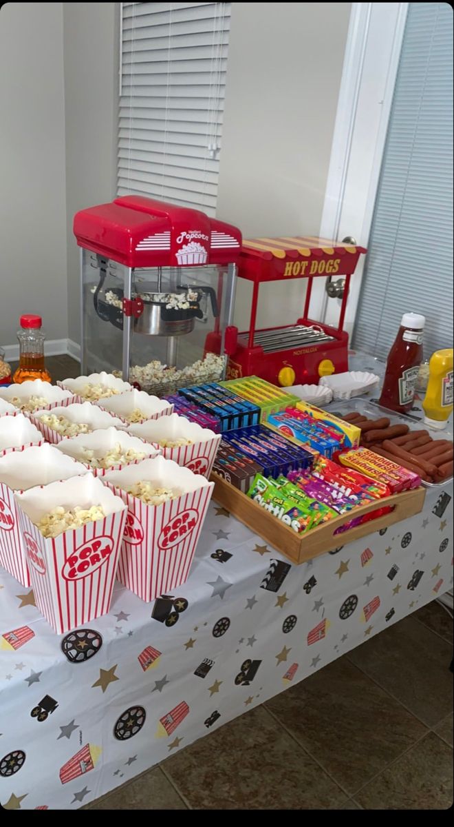 a table topped with popcorn buckets filled with cake and candy next to a hot dog stand