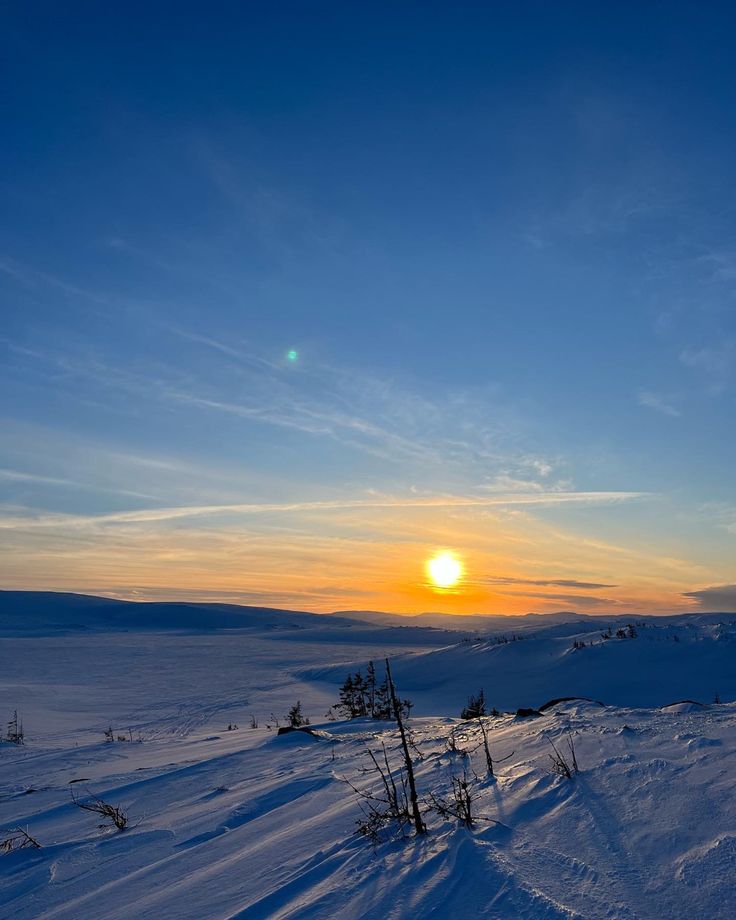 the sun is setting over some snow covered hills with trees in the foreground and blue skies above