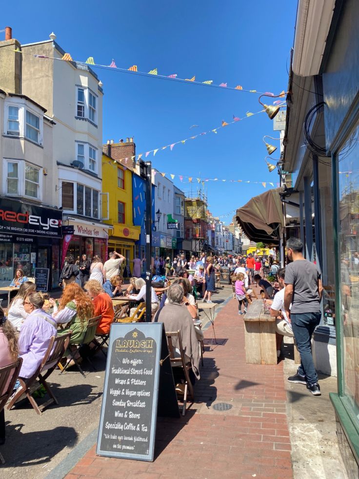 people are sitting at tables outside on the sidewalk in front of shops and buildings with bunting